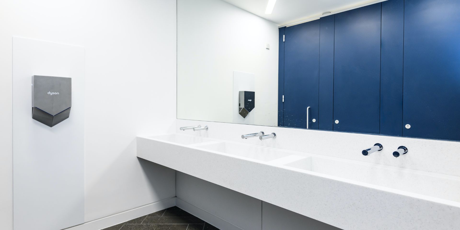 Sinks in a public toilet with a large wall length mirror above, and a Dyson hand dryer on the wall.